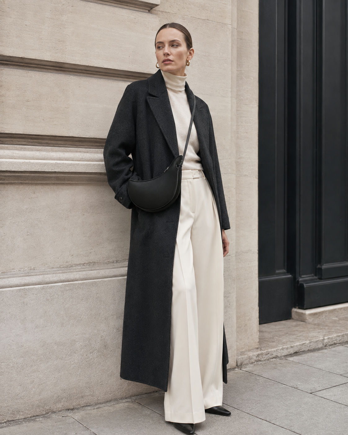 Woman in tailored neutrals carries the black MUYLOTTA ML-CB03 crescent crossbody bag by a limestone wall.