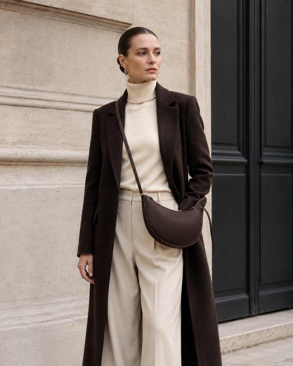 Woman in tailored neutrals carries the deep brown MUYLOTTA ML-CB03 crescent crossbody bag by a limestone wall.