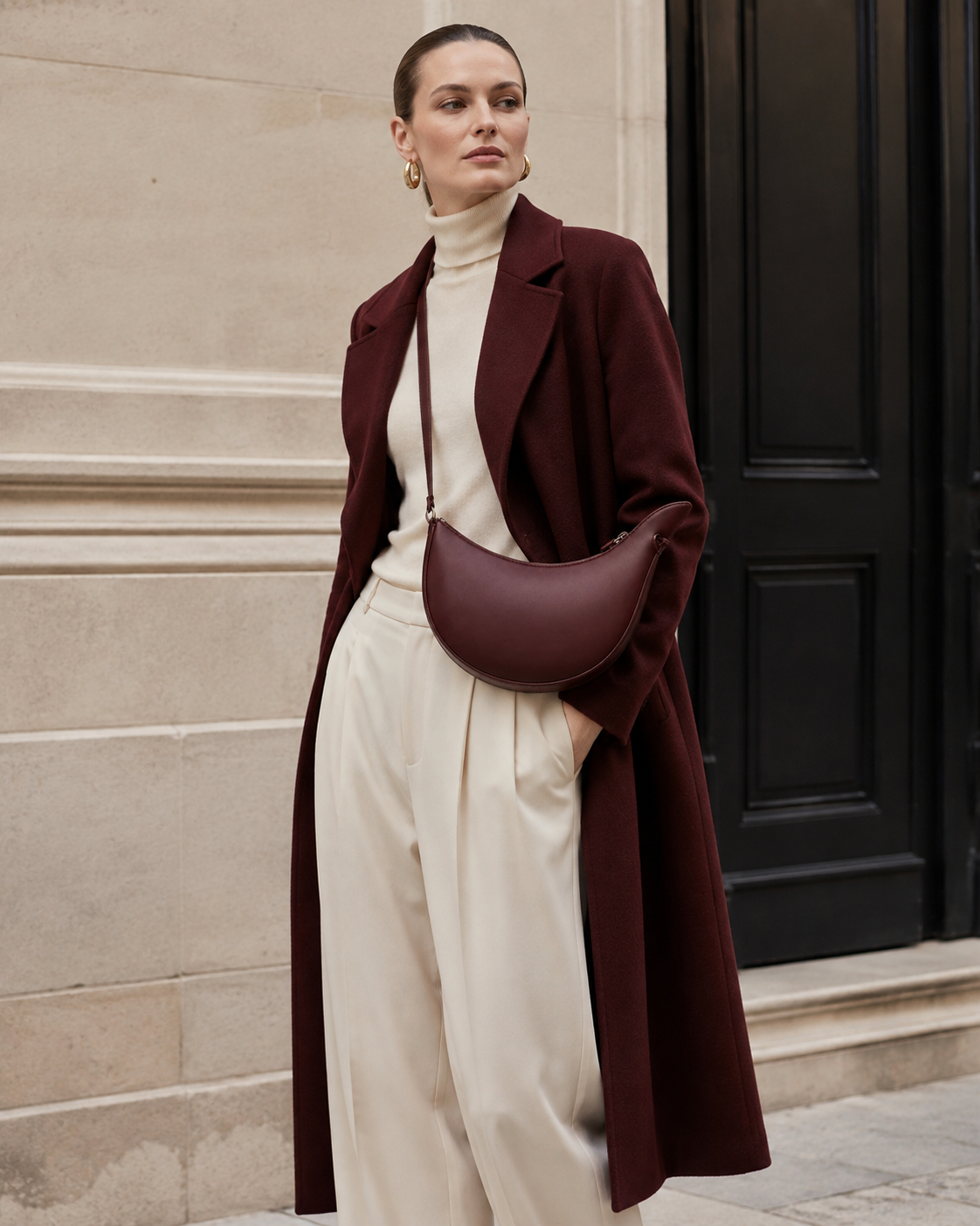 Woman in tailored neutrals carries the oxblood MUYLOTTA ML-CB03 crescent crossbody bag by a limestone wall.