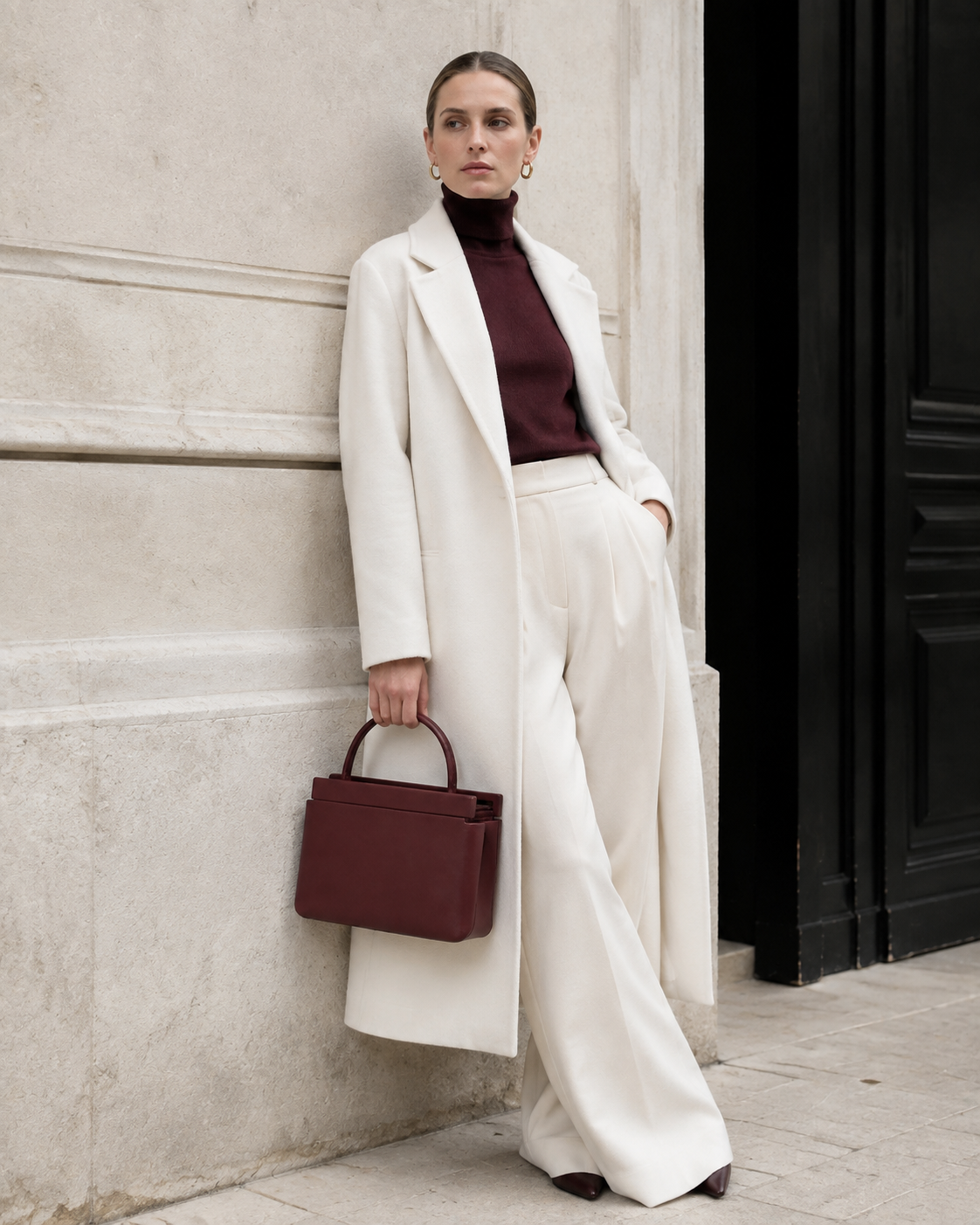 Woman in tailored neutrals carries the oxblood MUYLOTTA ML-MN01 mini top-handle bag by its top handle by a limestone wall.